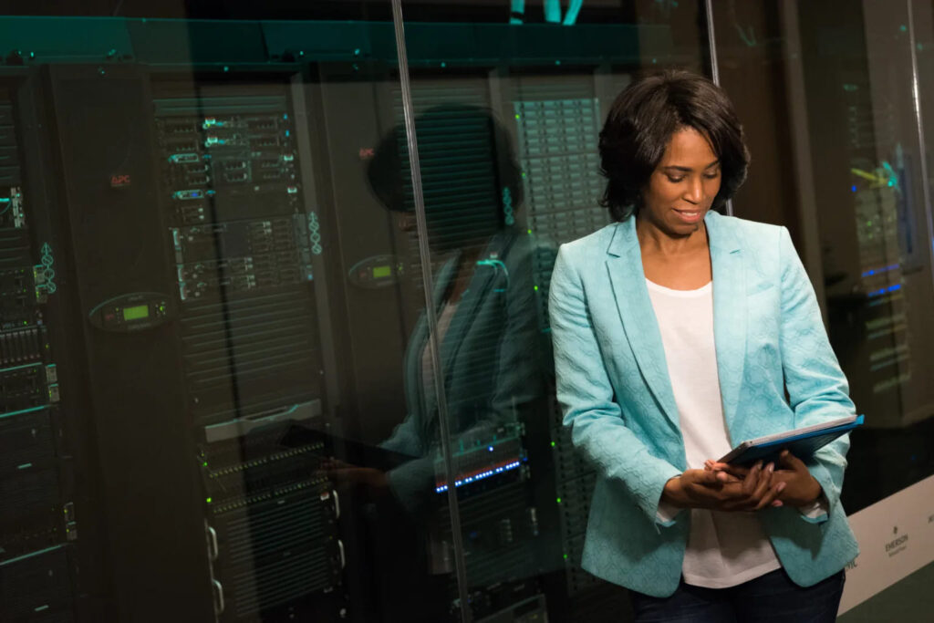 A woman looks at a tablet, standing next to tower servers