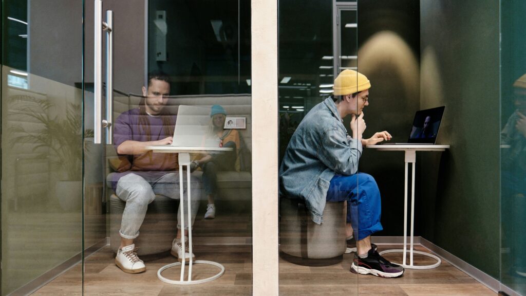 Two people working on laptops inside adjacent private glass office booths.