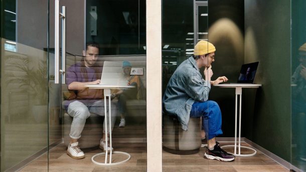 Two people working on laptops inside adjacent private glass office booths.
